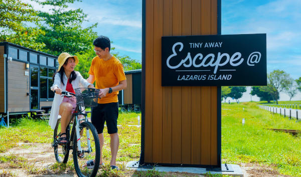 Couple with bikes at "Escape @ Lazarus Island" sign. Tiny house in background.